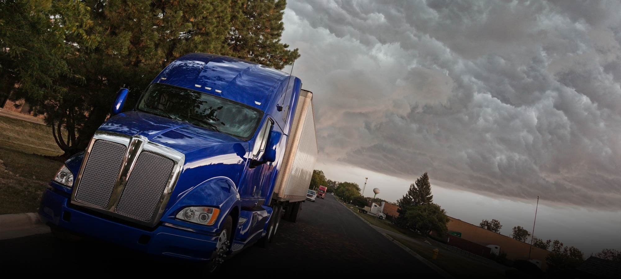 Tilted photo of a royal blue semi tractor and dry van parked along the side of the street with storm clouds building in the background