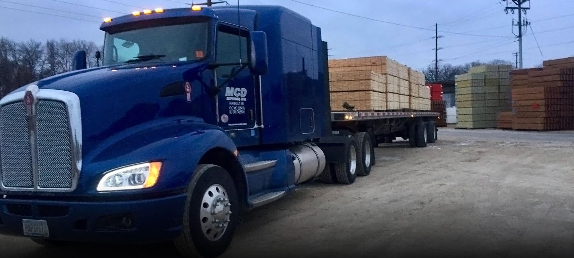 Royal blue MCD Express semi tractor connected to a flatbed trailer that's getting loaded with plywood and lumber